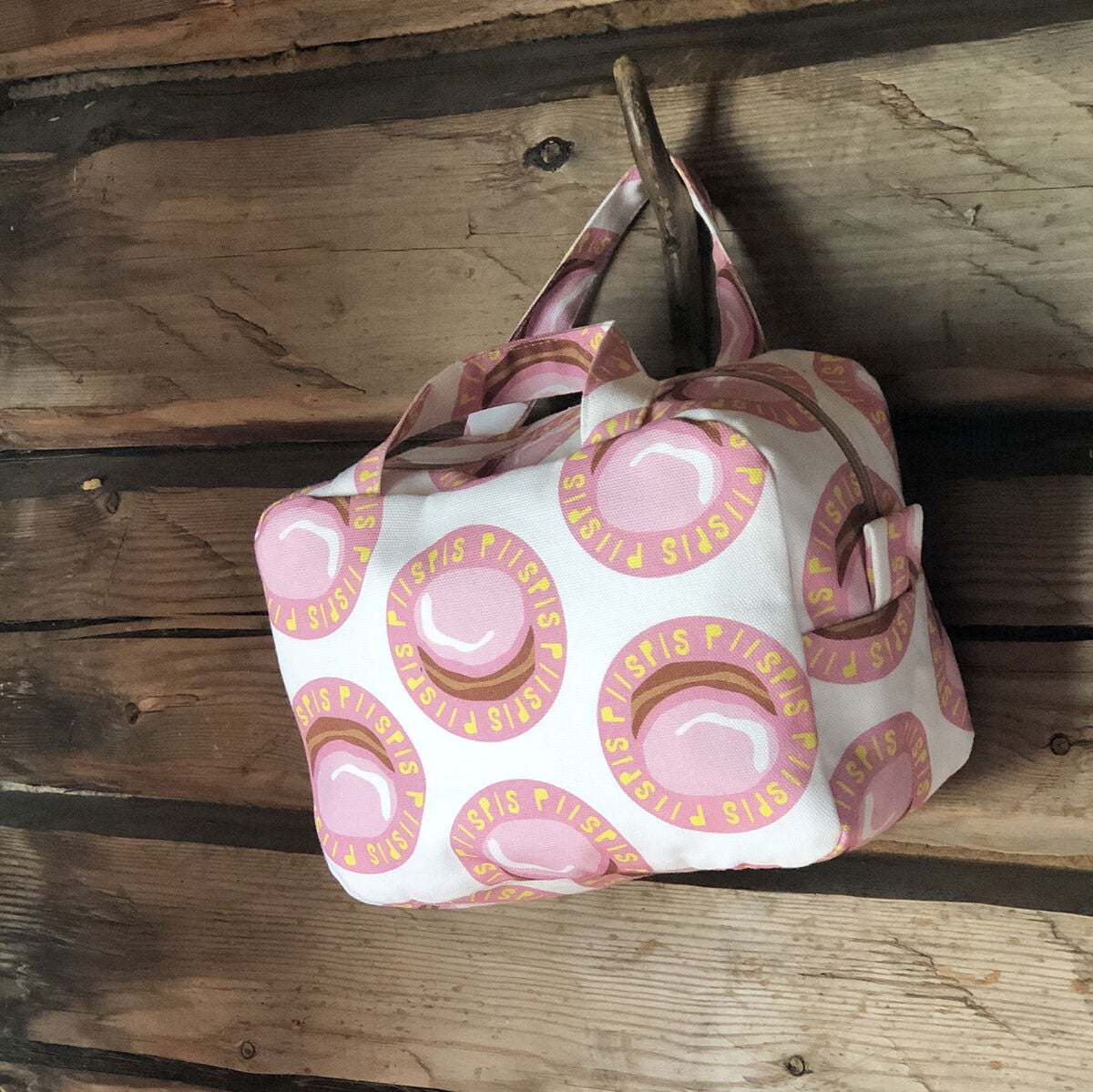 Kui designin Piispis-toilettilaukku roikkumassa puisesta seinäkoukusta. Valkoinen pohjaväri, jossa Piispanmunkki kuosi.